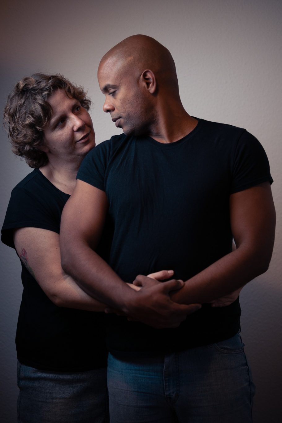 Pareja abrazándose, mirándose con ternura, ambos con camisetas negras.