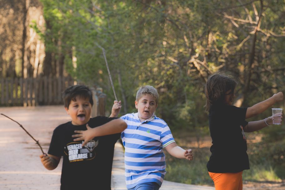 Tres niños juegan al aire libre, uno sonríe mientras los otros corren y sostienen palos.