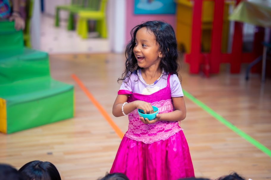 Niña con vestido rosa sonriendo y sosteniendo un objeto en un aula colorida.