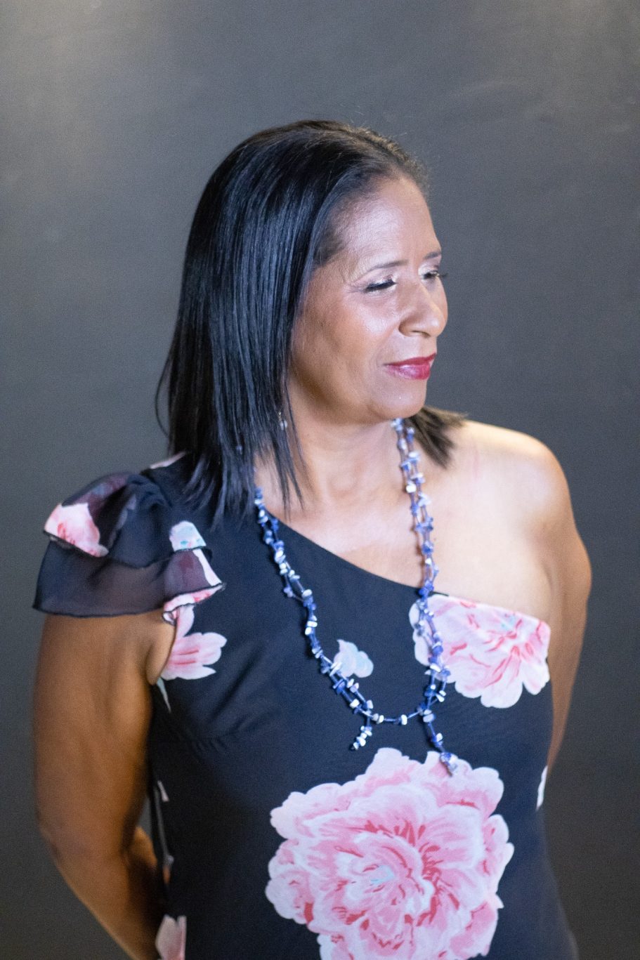 Mujer de pie con blusa floral y collar, mirando hacia un lado con expresión serena.