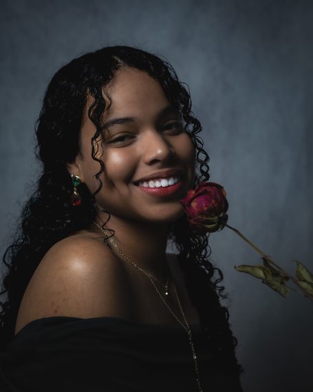 Mujer sonriente con cabello rizado sostiene una rosa encarnada cerca de su rostro.