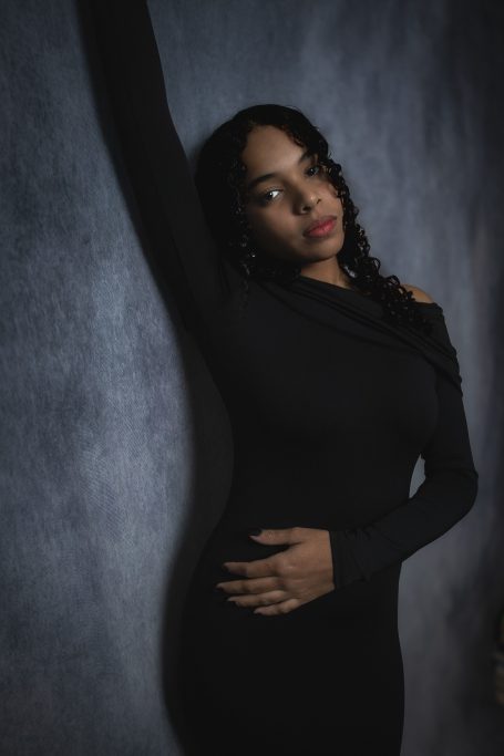 Mujer con vestido negro, posando contra un fondo azul oscuro, con una expresión seria.