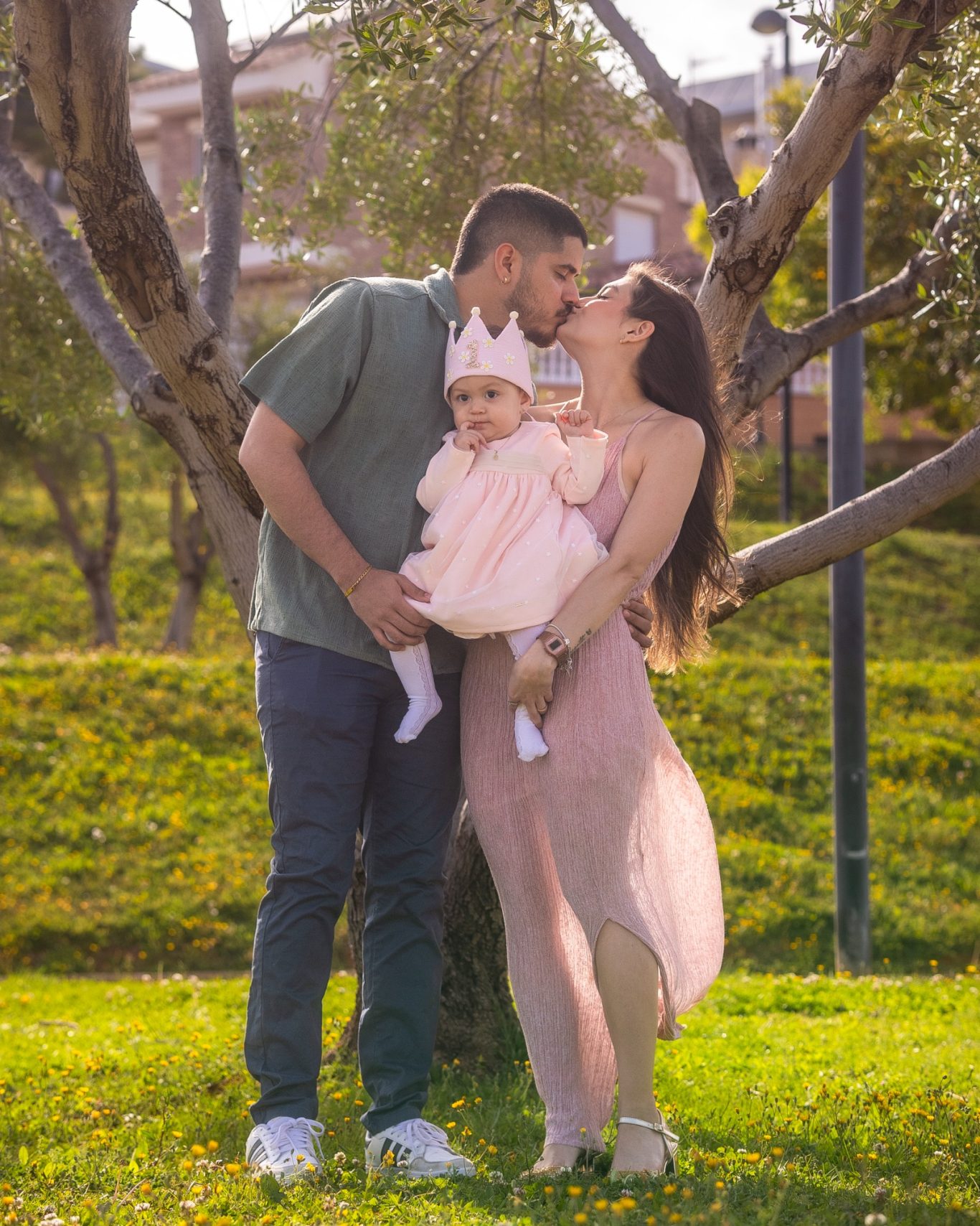 Familia feliz de pie al aire libre, con un padre, madre y su bebé en un vestido rosa.