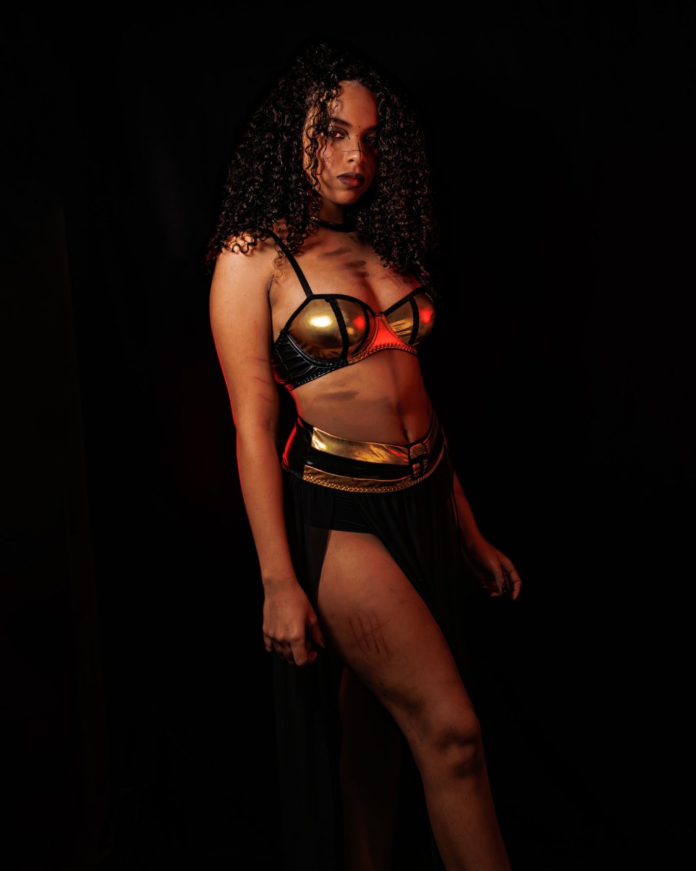 Mujer con cabello rizado, vestida con un conjunto oscuro y dorado, posando en un fondo negro.