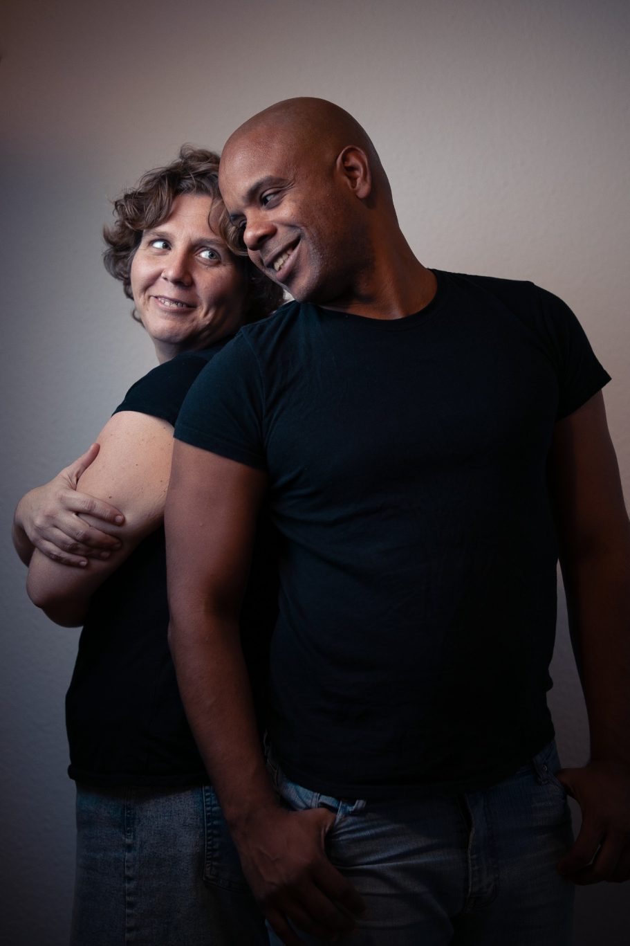 Pareja de personas sonriendo, de pie espalda con espalda, usando camisetas de manga corta negras.