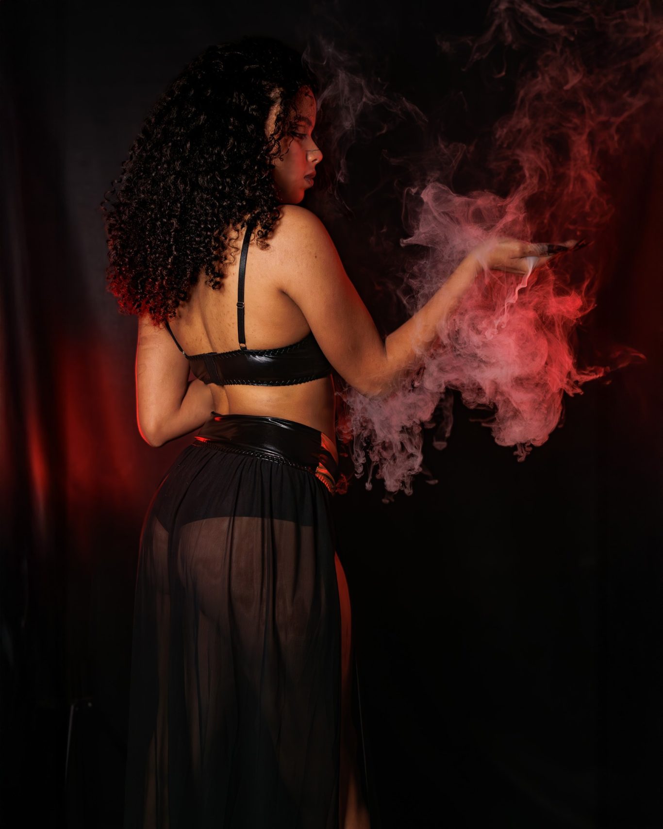 Mujer con cabello rizado, viste lencería negra y sostiene un cigarrillo entre nébulas de humo.
