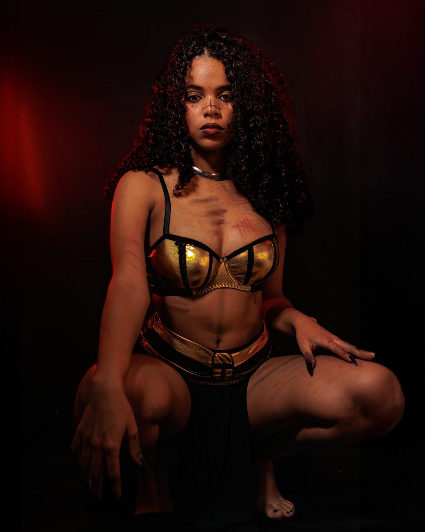 Persona con cabello rizado, vestida con un sujetador dorado y una falda negra, en un fondo oscuro.