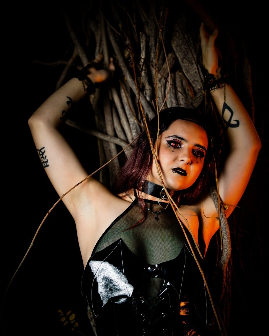 Joven con maquillaje oscuro y corsé, rodeada de ramas en un entorno sombrío.
