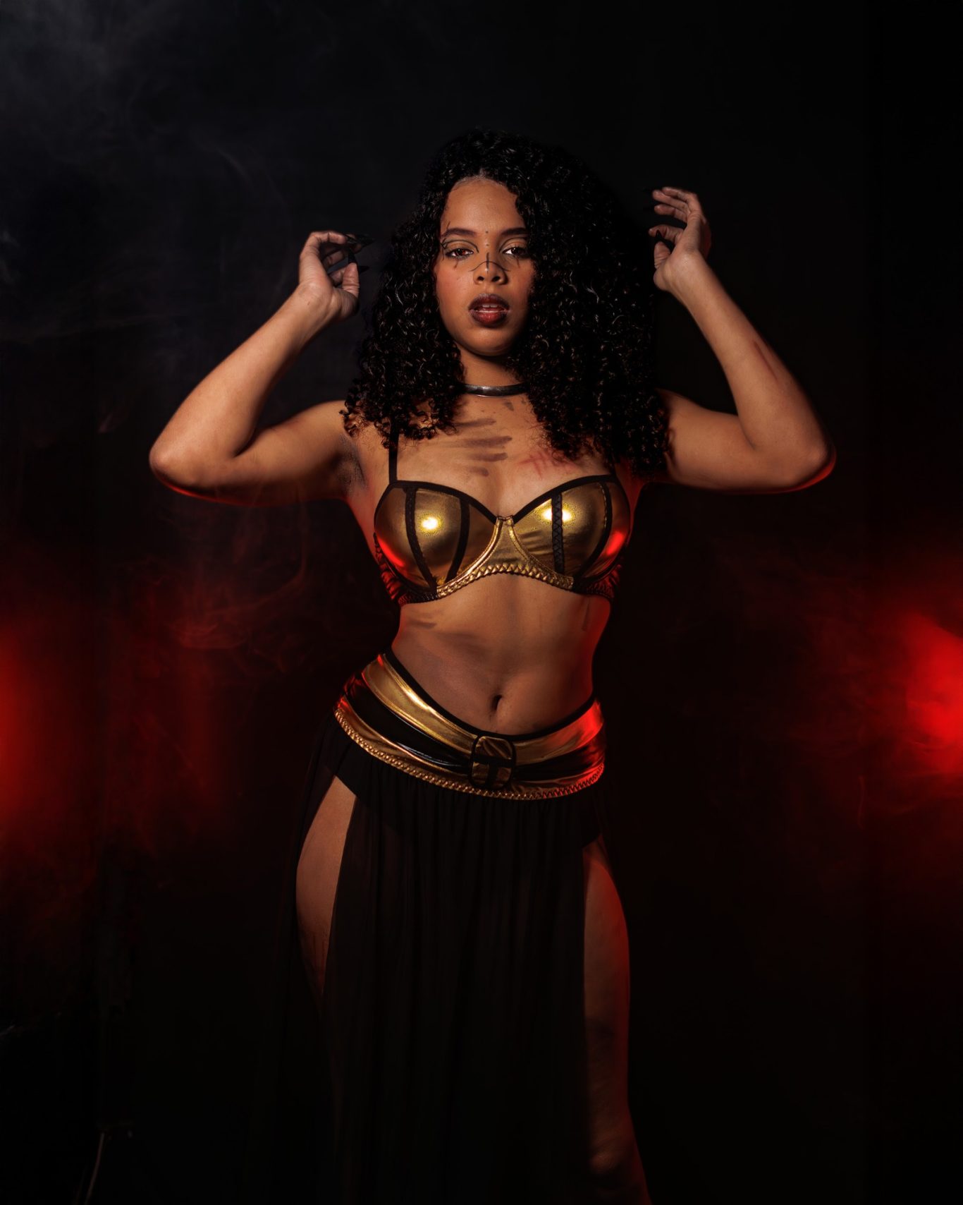 Mujer con cabello rizado, vestida con un top dorado y falda negra, en un fondo oscuro.