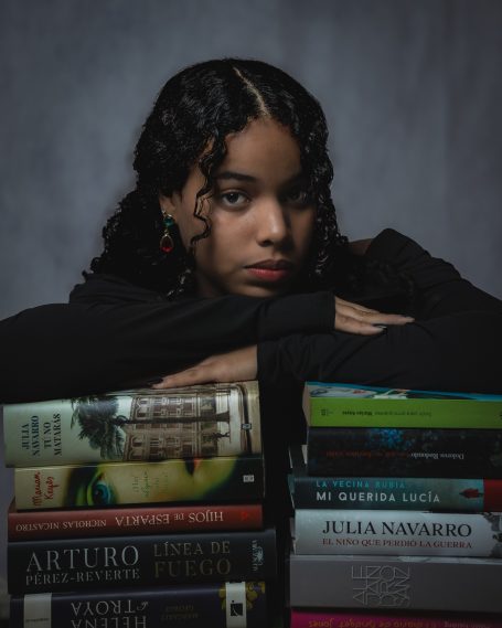 Joven con cabello rizado, apoyada sobre una pila de libros, mirando hacia adelante.