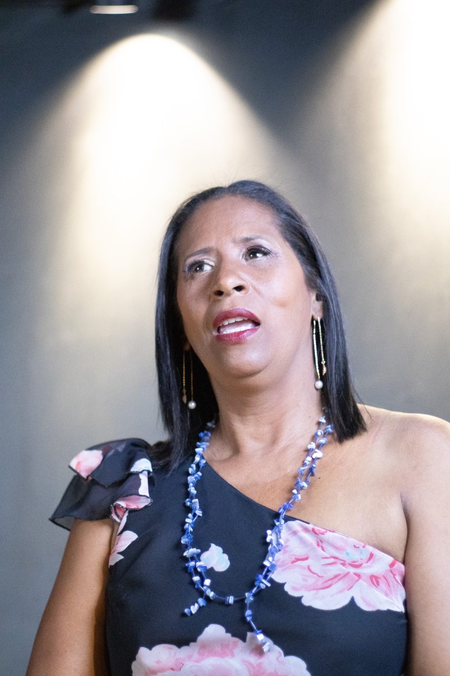 Mujer de pie, con blusa de flores y collar, mirando hacia arriba en un ambiente iluminado.
