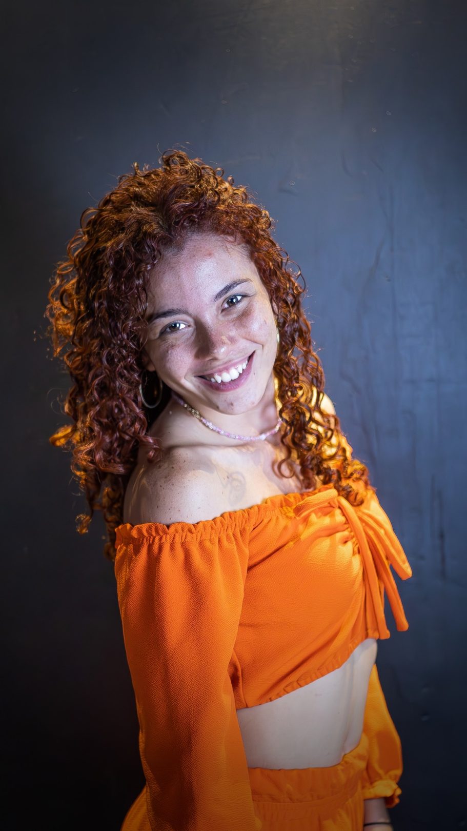 Mujer sonriente con cabello rizado, vistiendo un top naranja en fondo negro.