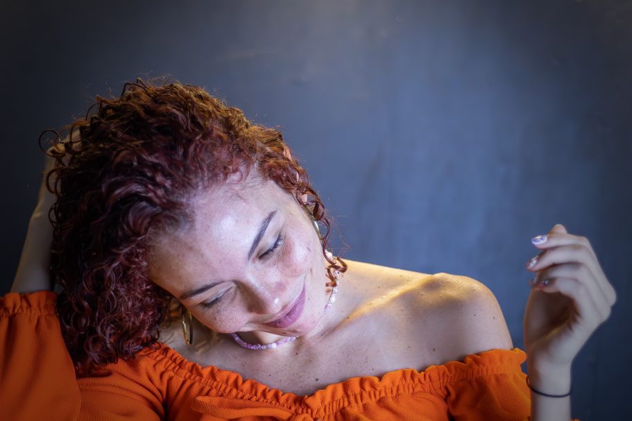 Mujer con cabello rizado y rojizo, vistiendo una blusa naranja, mirada reflexiva.