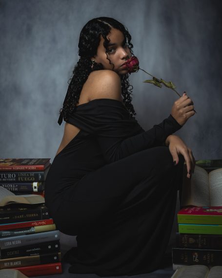 Mujer con cabello rizado y rosa en la boca, sentada junto a pilas de libros.