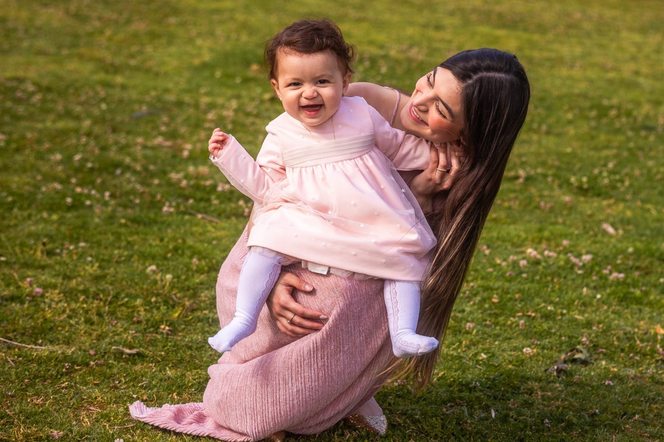 Mujer con cabello largo sostiene a una niña sonriente sobre su cadera en un jardín.