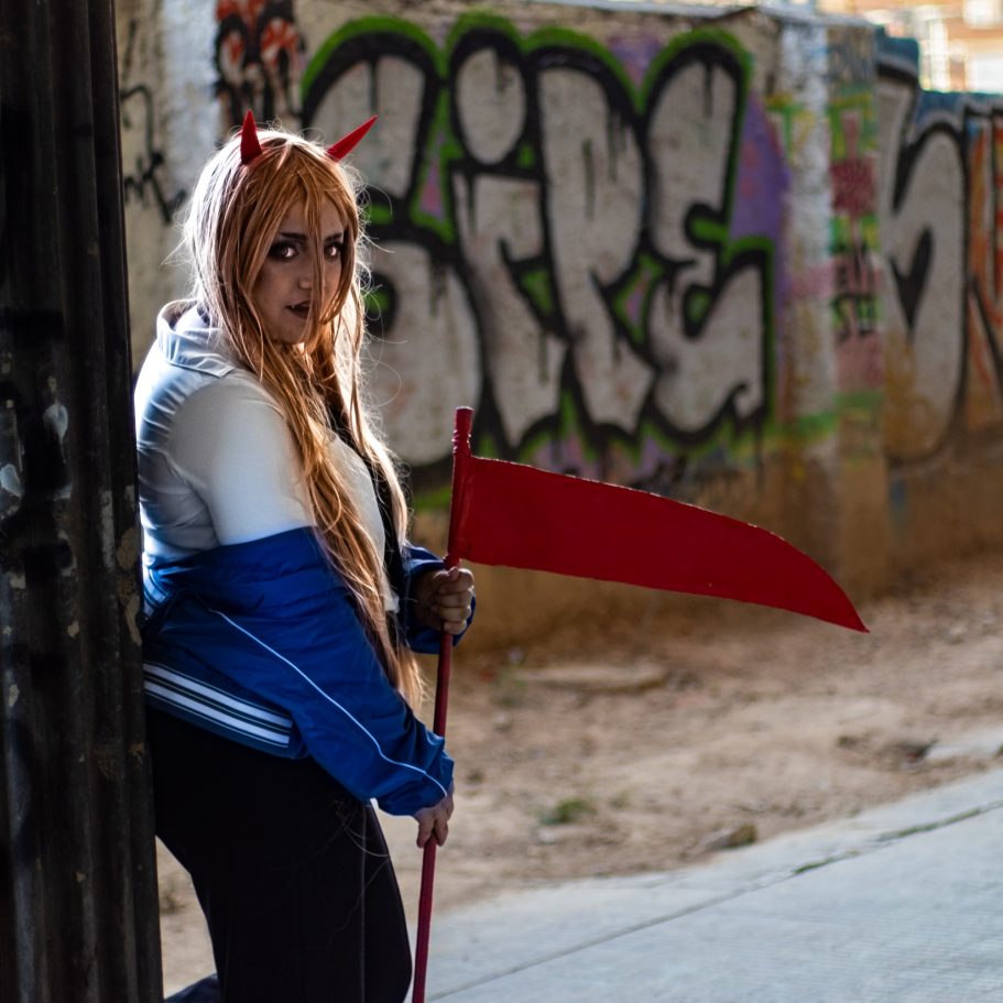 Persona con cuernos y cabello largo, vestida con chaqueta azul, sosteniendo una guadaña roja.