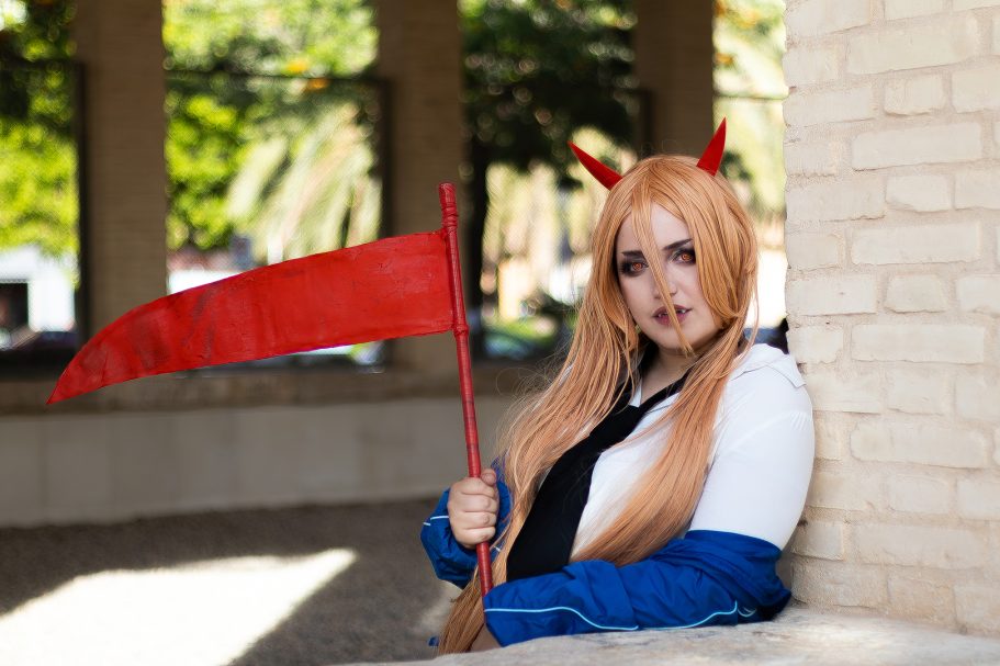 Cosplay de persona con cabello rubio y bandera roja, fondo de árboles y columnas.