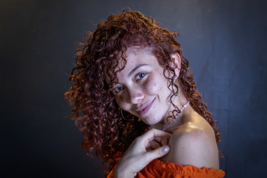 Mujer con cabello rizado y rojizo, sonriendo, con fondo oscuro.