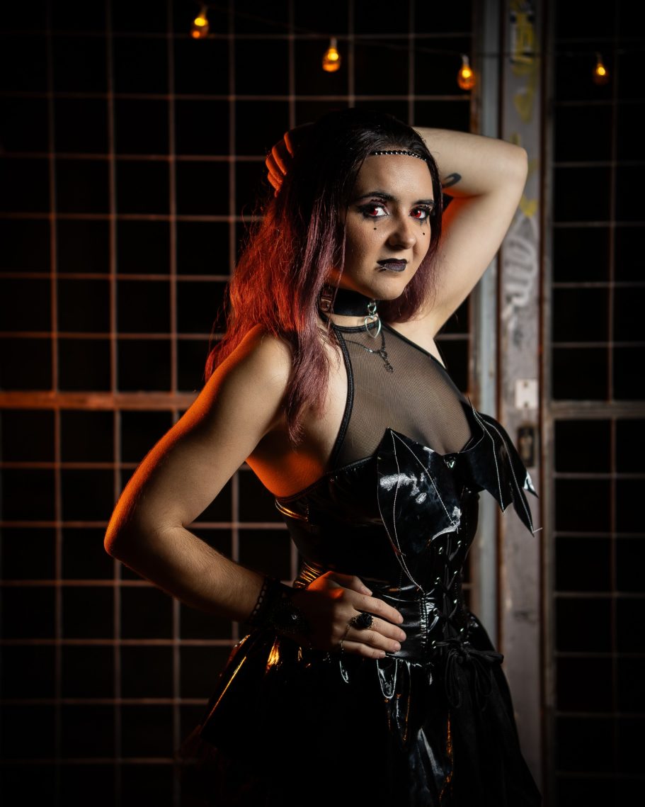 Mujer con cabello largo y morado, vistiendo un corsé negro y posando en un entorno oscuro.