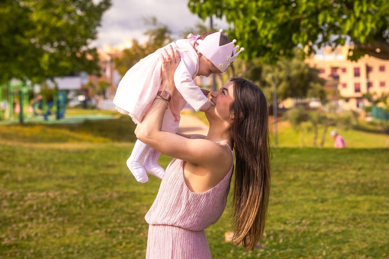 Mujer sostiene a un bebé en un parque, con árboles y juegos de fondo.