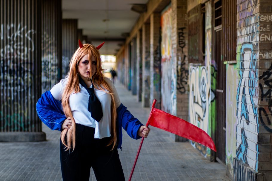 Persona con peluca larga y roja, vestida con una camisa blanca y pantalones negros, sosteniendo una bandera roja.