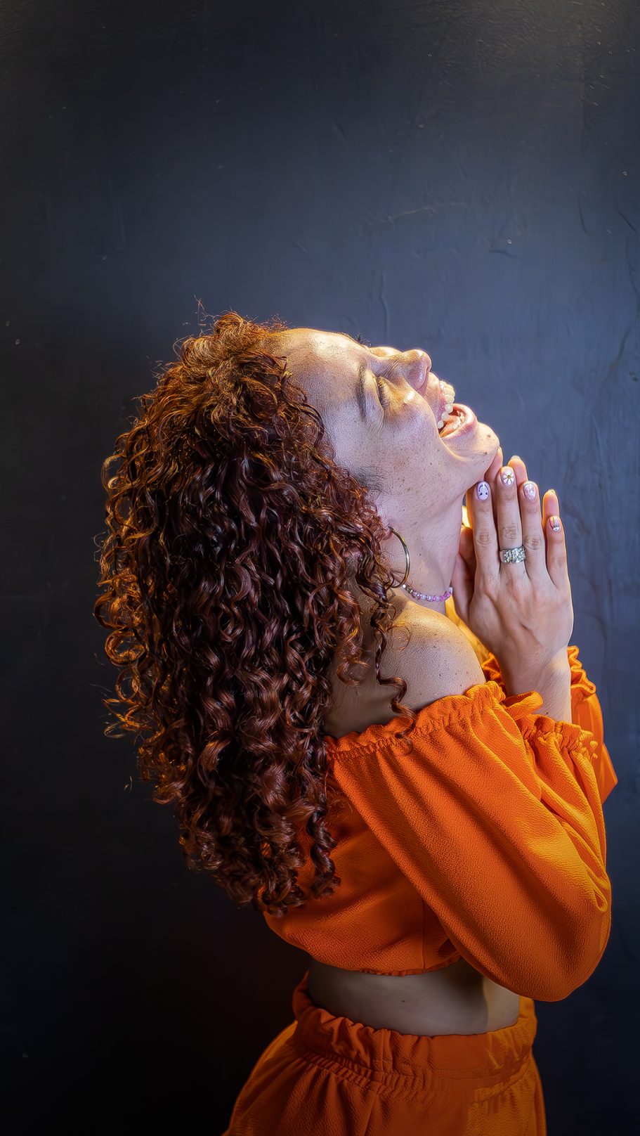 Mujer con cabello rizado, vestida de naranja, mirando al cielo con las manos en oración.