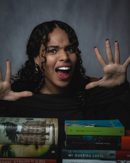 Mujer con cabello rizado, expresando sorpresa, rodeada de libros apilados.
