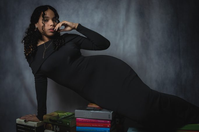 Mujer en vestido negro recostada sobre libros con fondo oscuro.