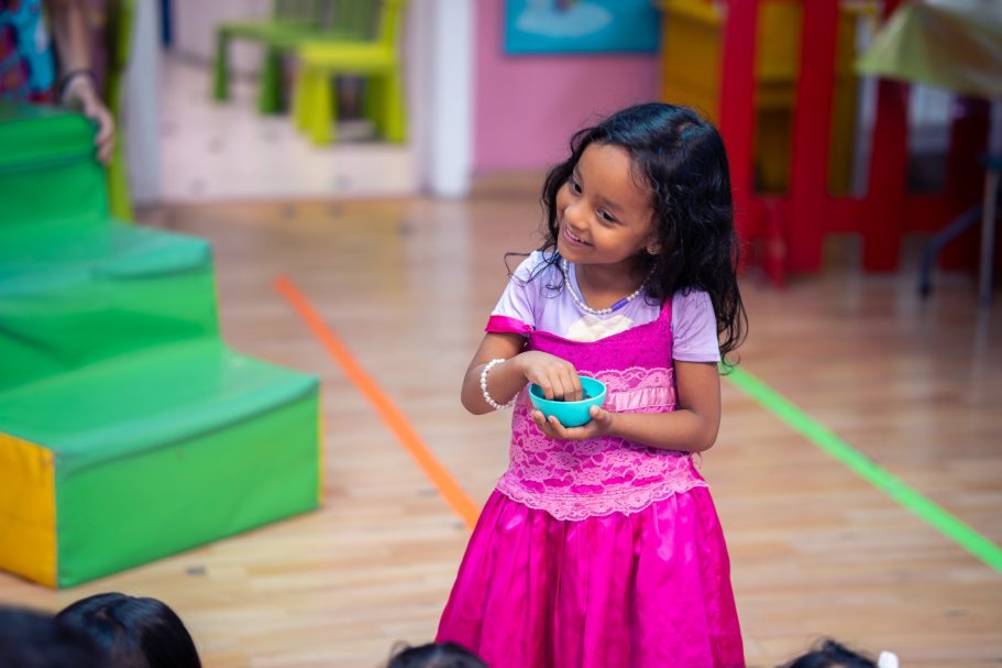 Niña con vestido rosa y con una pulsera, sonriendo mientras sostiene un objeto en la mano.