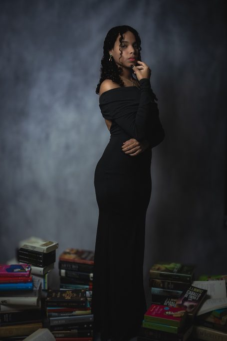Mujer de pie en un vestido negro, rodeada de libros apilados. Fondo gris.