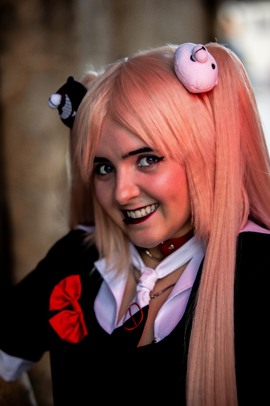 Persona con cabello largo y rosa, vestida con un uniforme negro y una cinta roja.