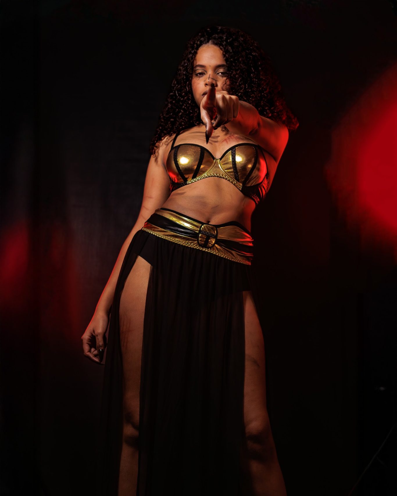 Mujer con vestuario dorado y negro, posando en un fondo oscuro y iluminado con luces rojas.