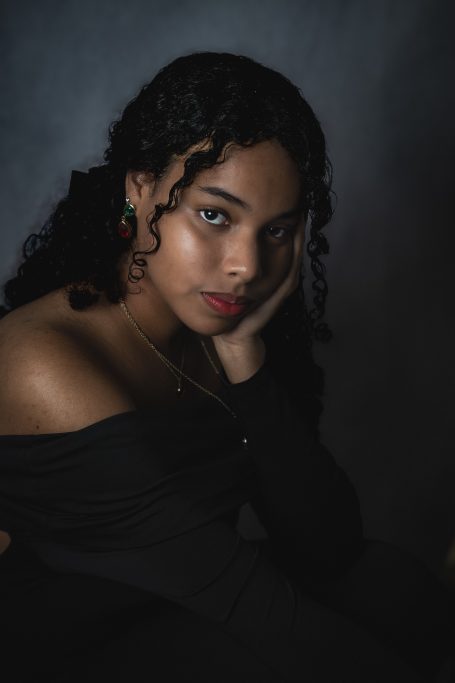Mujer con cabello rizado, posando con una mano en la cara, fondo oscuro y sutil iluminación.