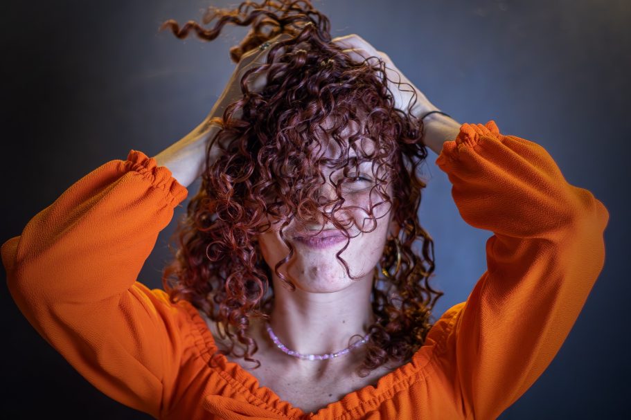Mujer con cabello rizado y desordenado, levantando las manos y vistiendo una blusa naranja.