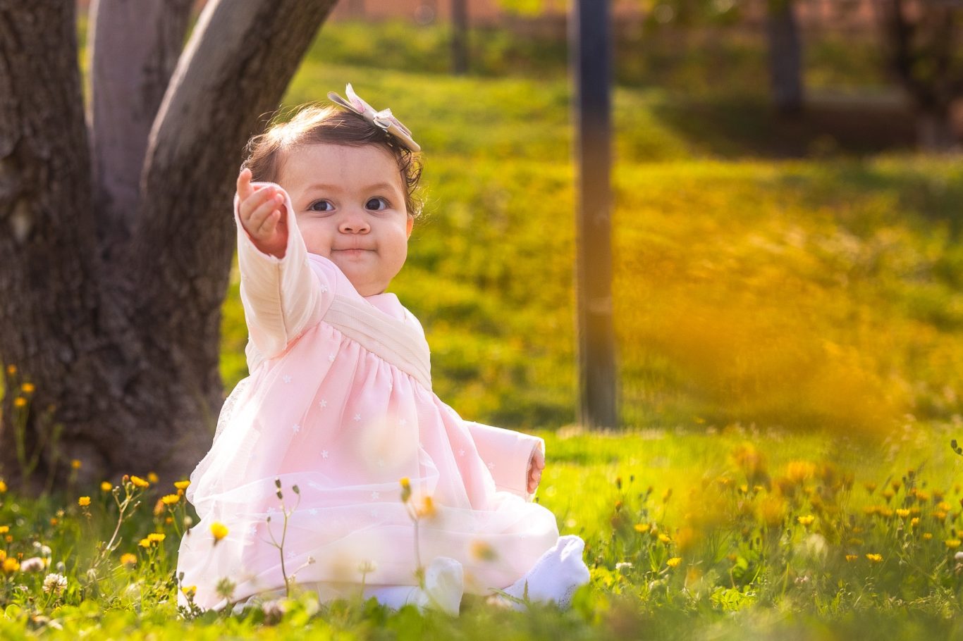 Niña pequeña sentada en el césped, señalando con una mano y sonriendo.